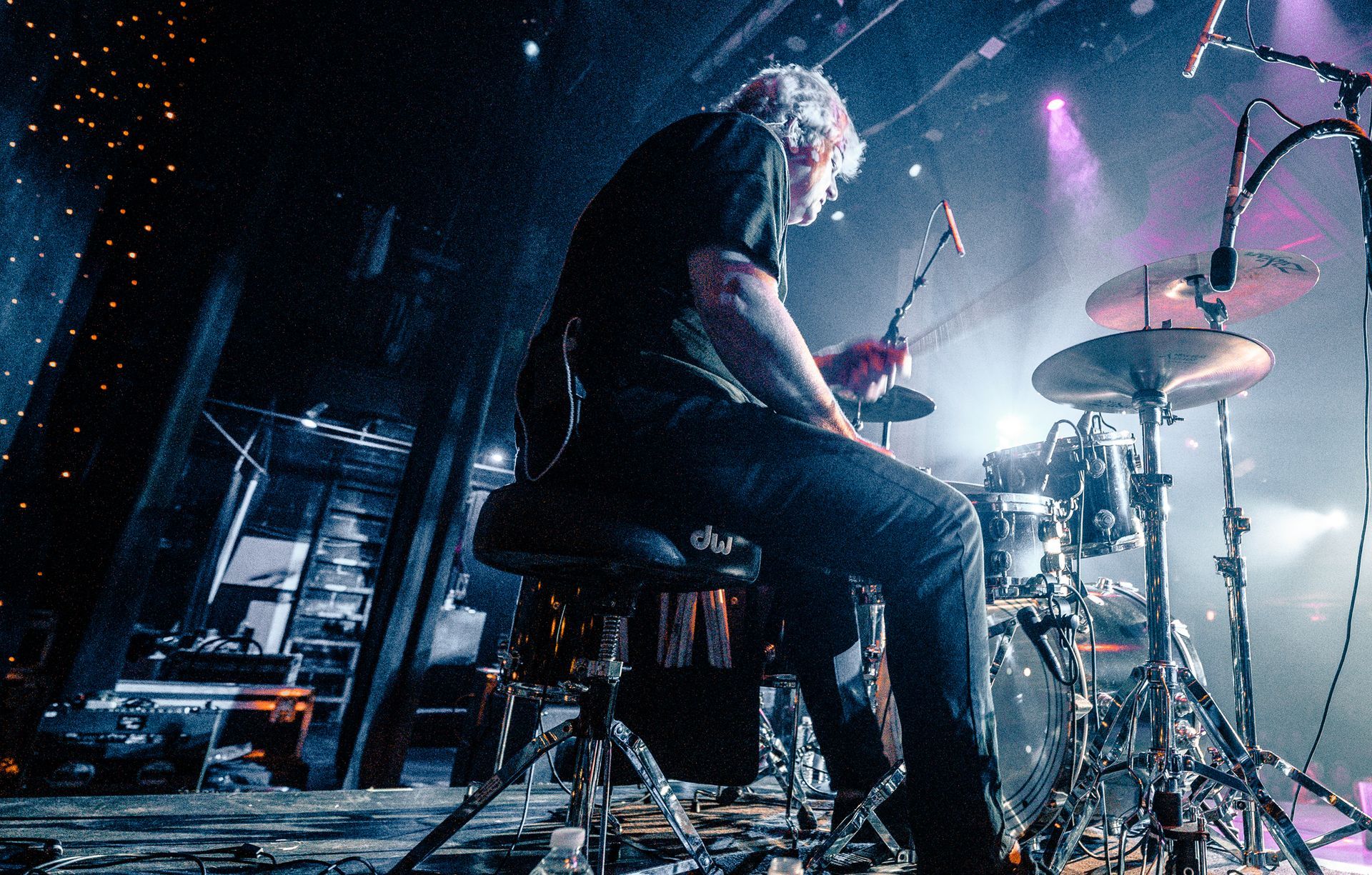 Drummer playing on stage, viewed from behind. Drum set, dark lighting, concert setting.
