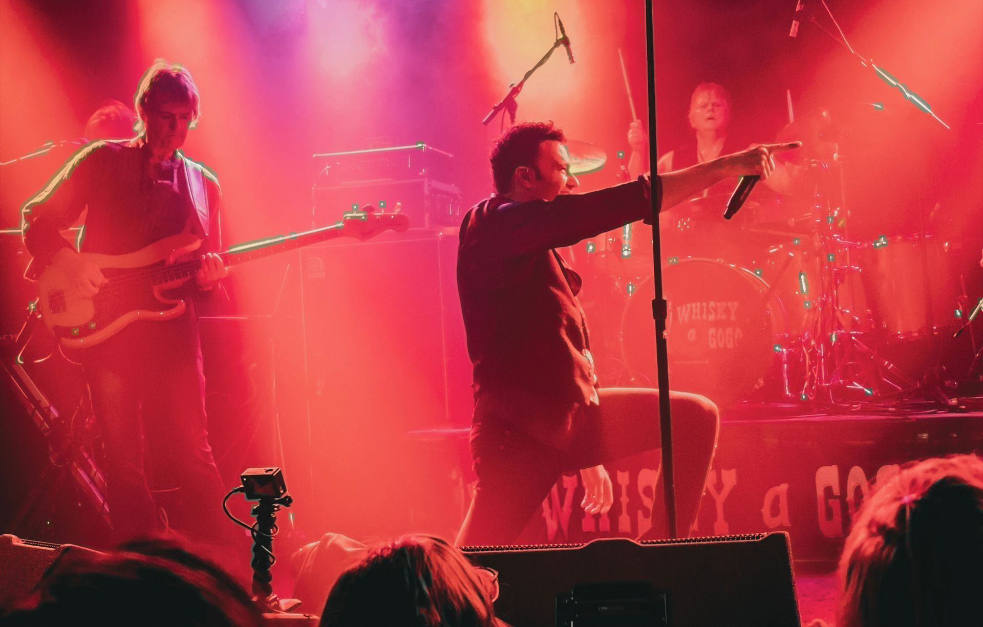 Band performing on stage, singer kneeling, pointing with microphone, bathed in red stage lighting.