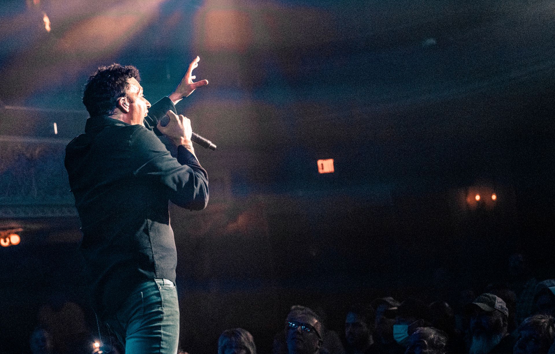 Man on stage, singing into a microphone, with arm raised. Crowd in the blurred foreground. Dim lighting.