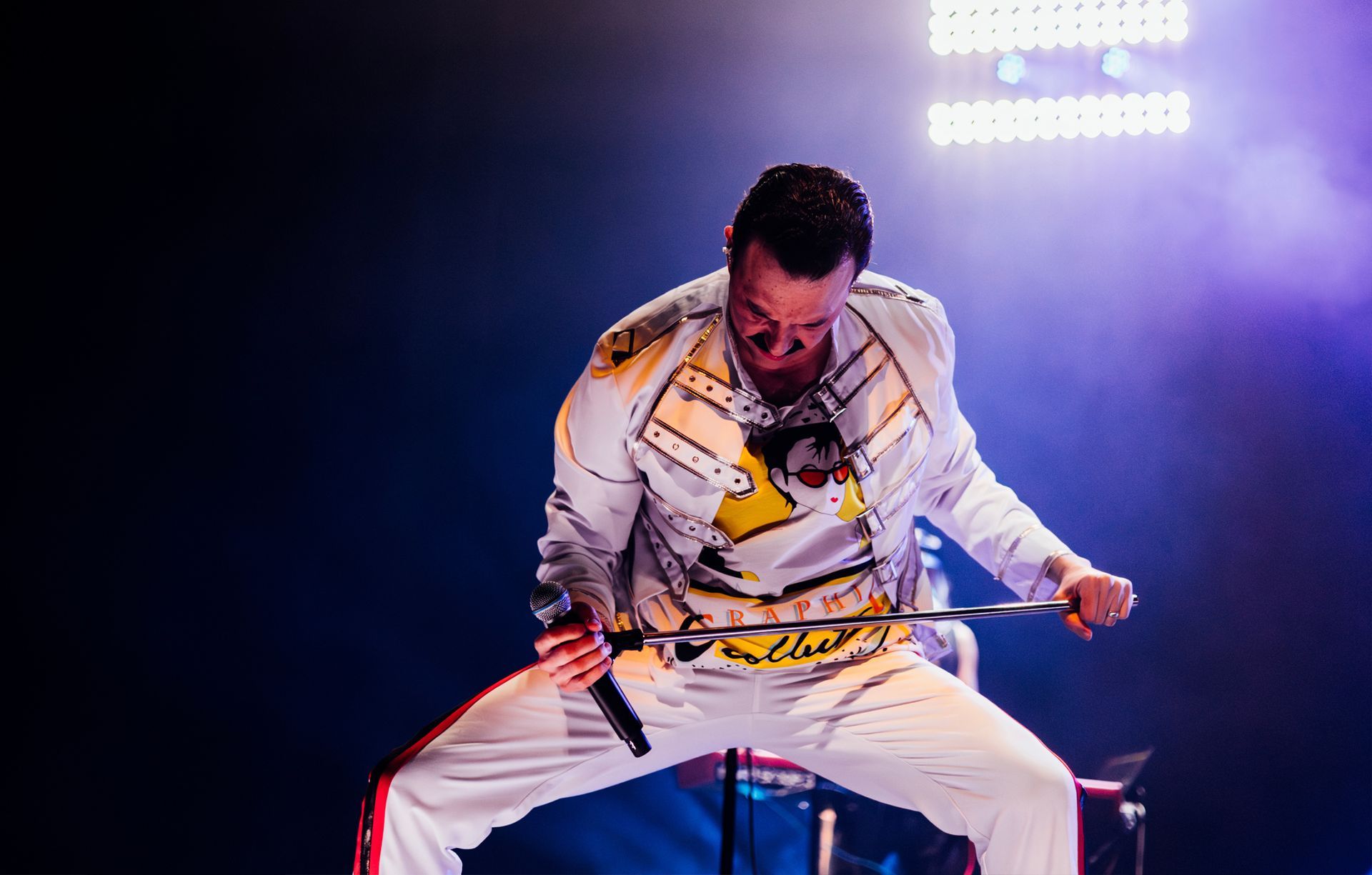 Man in white jacket, impersonating Freddie Mercury, singing on stage, holding microphone, dynamic pose.