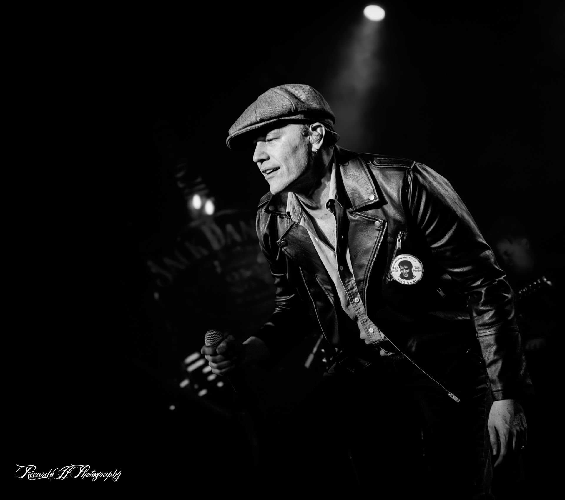 A man in a leather jacket and hat is singing into a microphone on a stage.