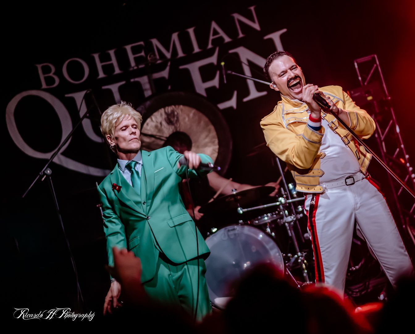 Two men in suits are singing into microphones on a stage in front of a crowd.