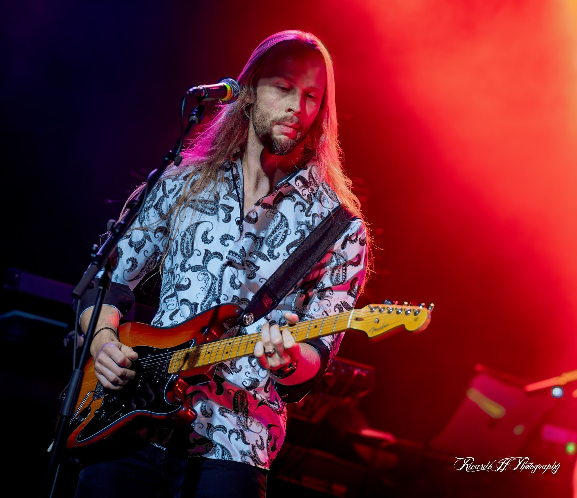 A man with long hair is playing a guitar and singing into a microphone on a stage.