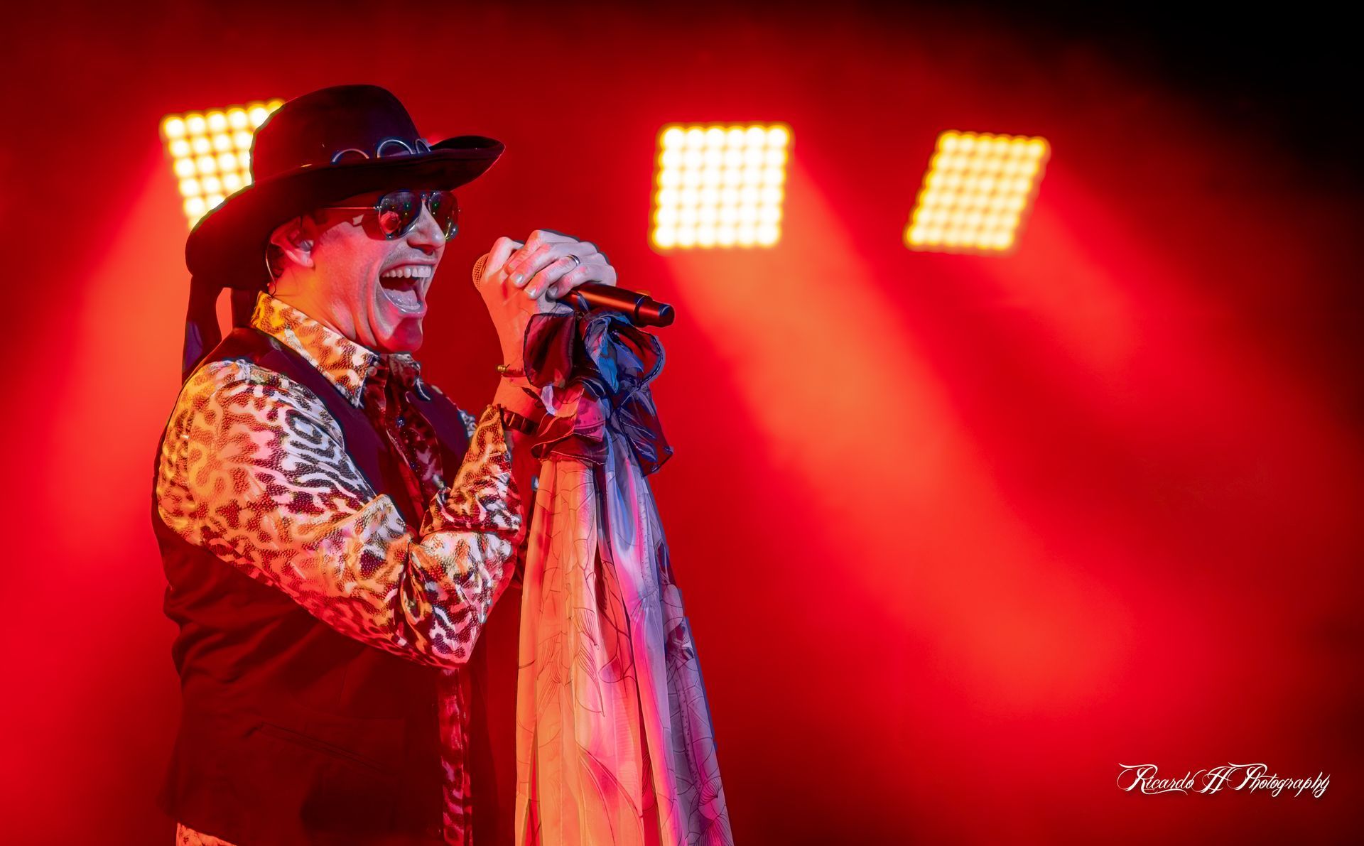 A man in a hat and sunglasses is singing into a microphone on a stage.