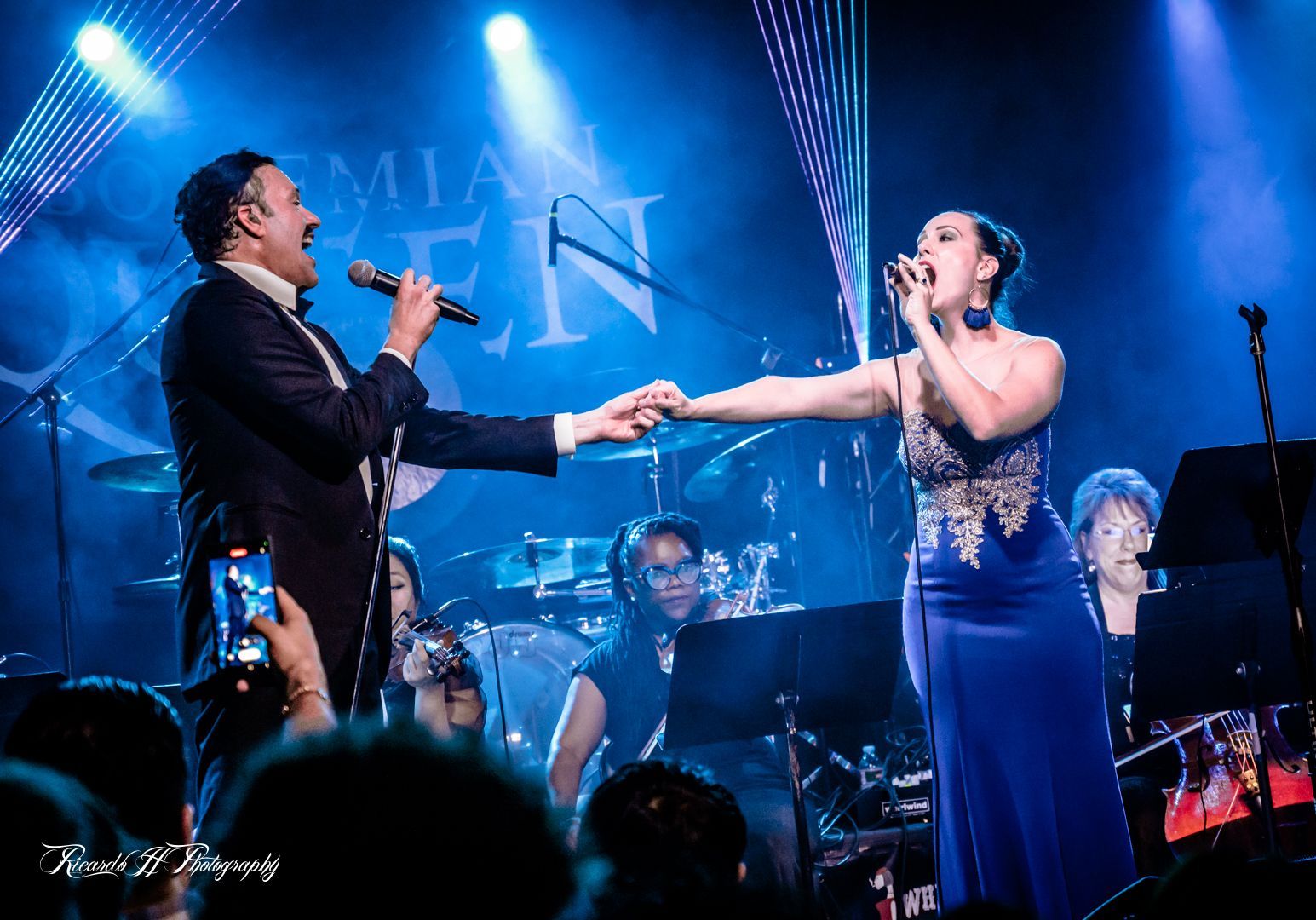 A man in a tuxedo and a woman in a blue dress are singing on a stage.