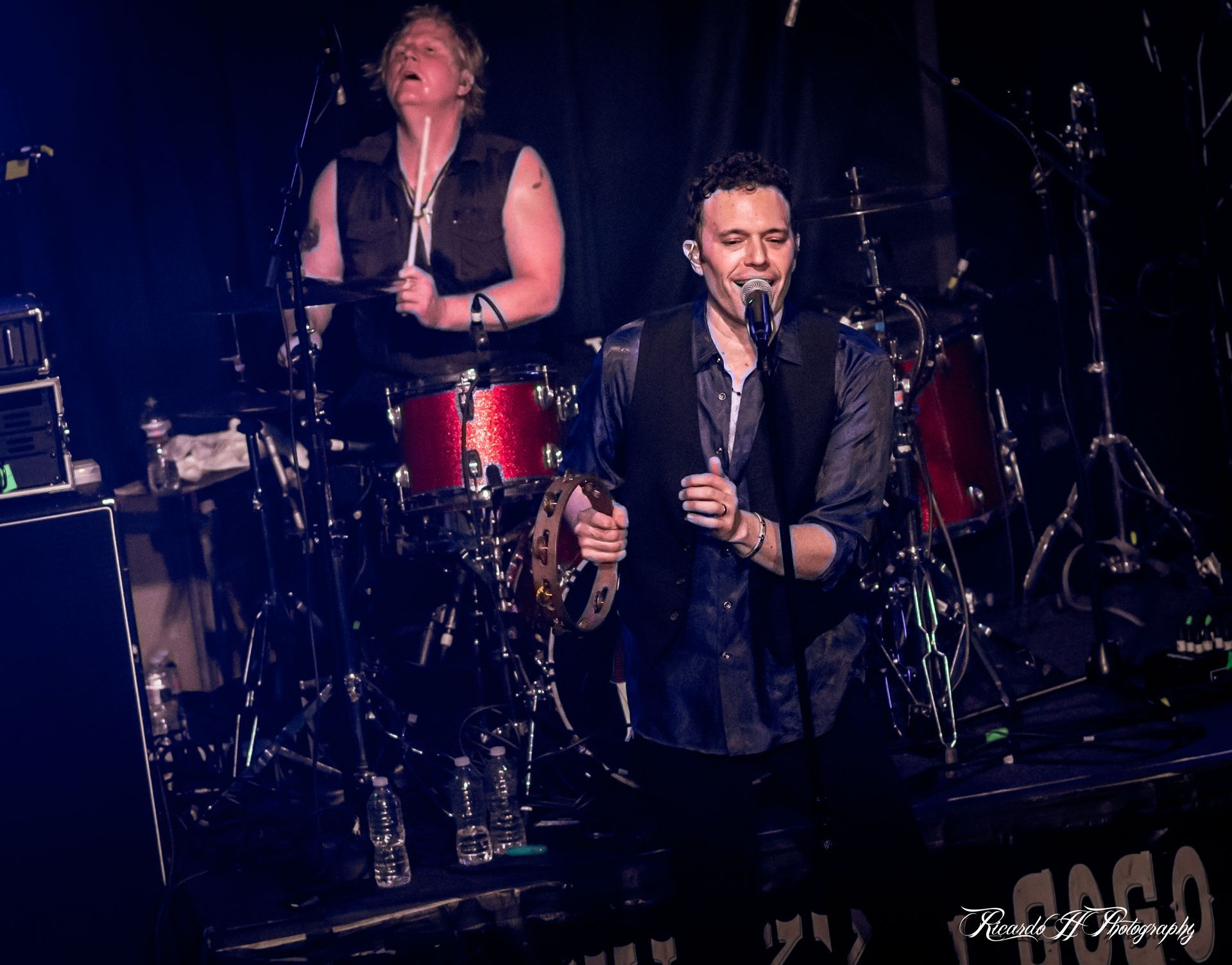 A man is singing into a microphone on a stage while another man plays drums.