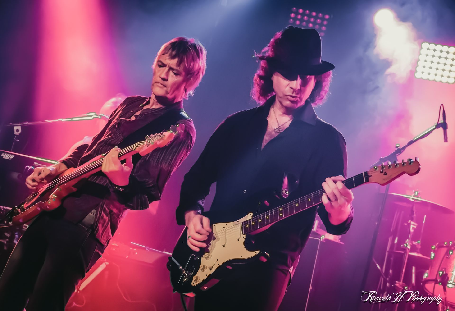 Two men are playing guitars on a stage in front of a microphone.