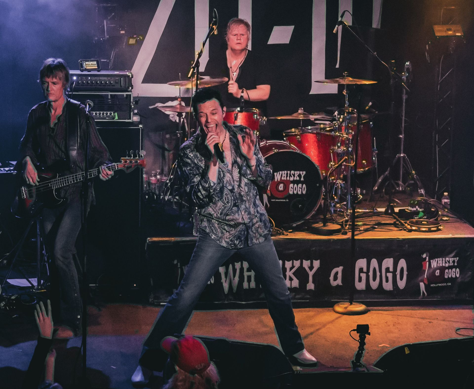 Band performing on stage at the Whisky a Go Go. Singer in a patterned shirt, guitarist, and drummer in background.