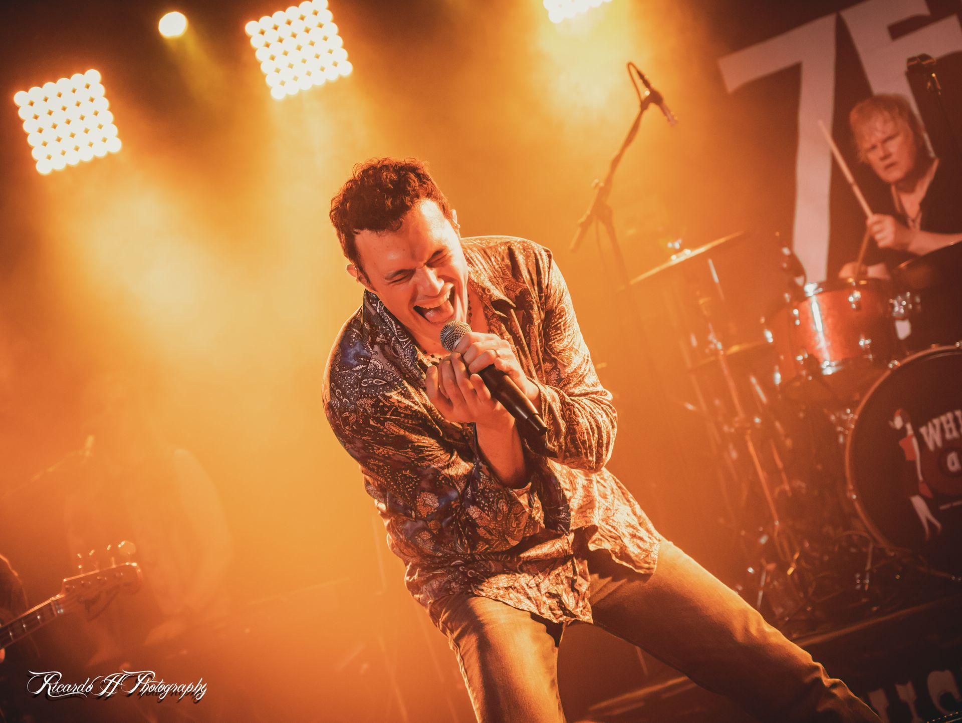 Singer on stage, singing into a microphone, bathed in orange light, drummer in background.