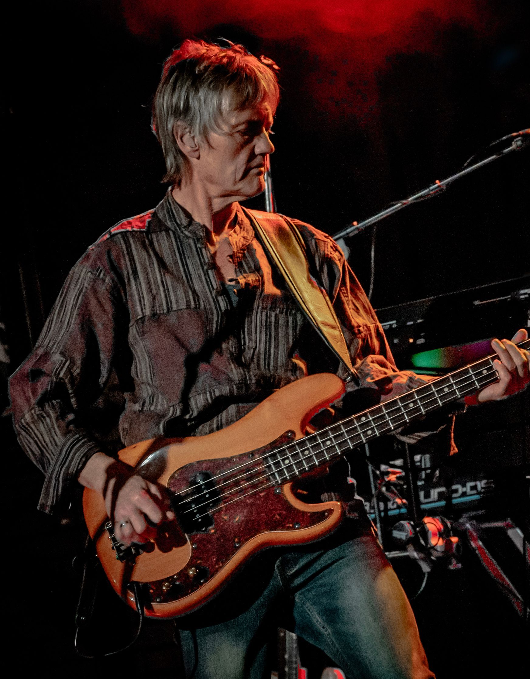 Man playing a brown bass guitar onstage, wearing a patterned shirt and jeans, under stage lighting.