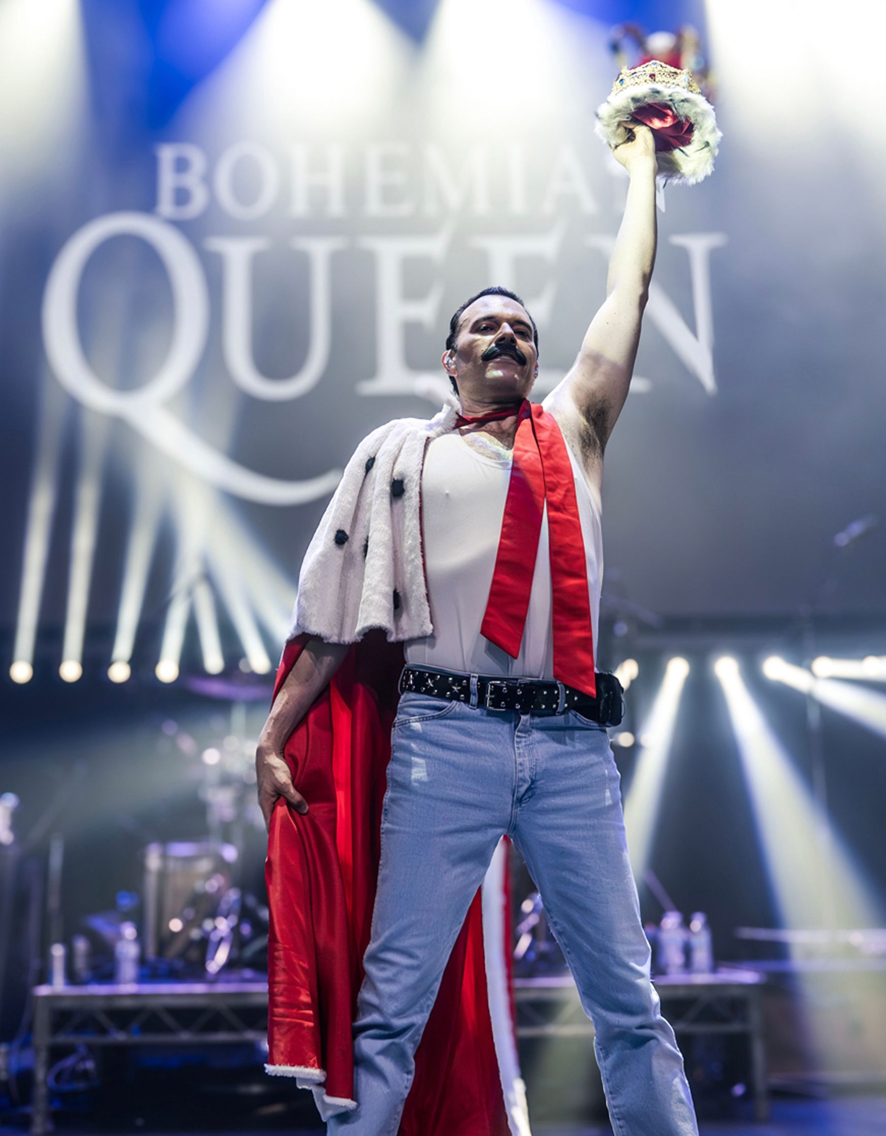 Man dressed as Freddie Mercury on stage, holding a crown, performing with a red cape.