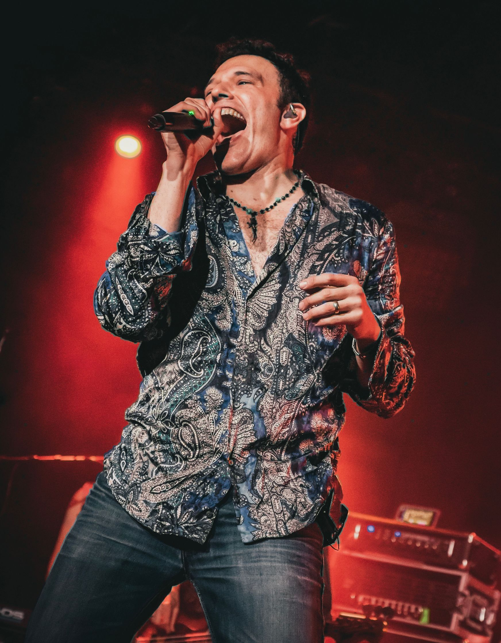 Male singer, microphone in hand, singing on stage with a patterned shirt and jeans. Red stage lighting.