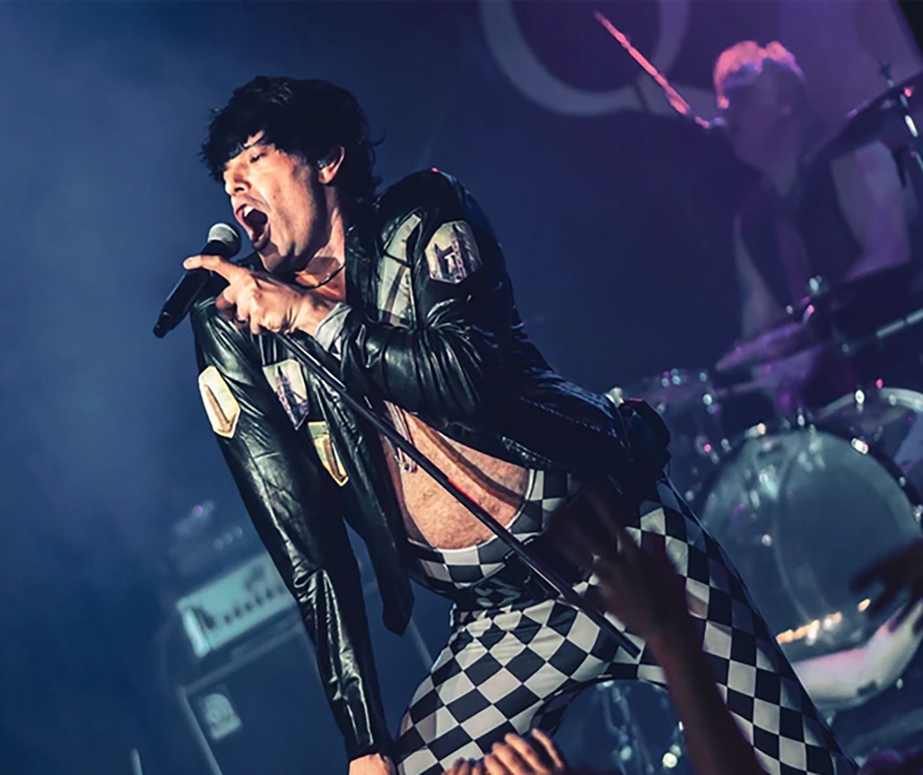 Singer on stage wearing leather jacket and checkered pants, singing into a microphone.