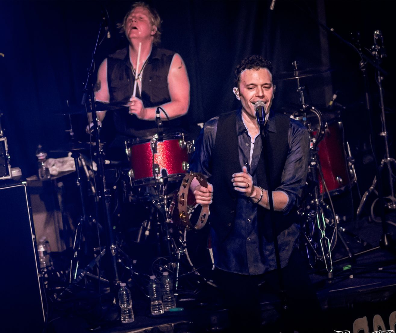 A man is singing into a microphone while another man plays drums on stage.