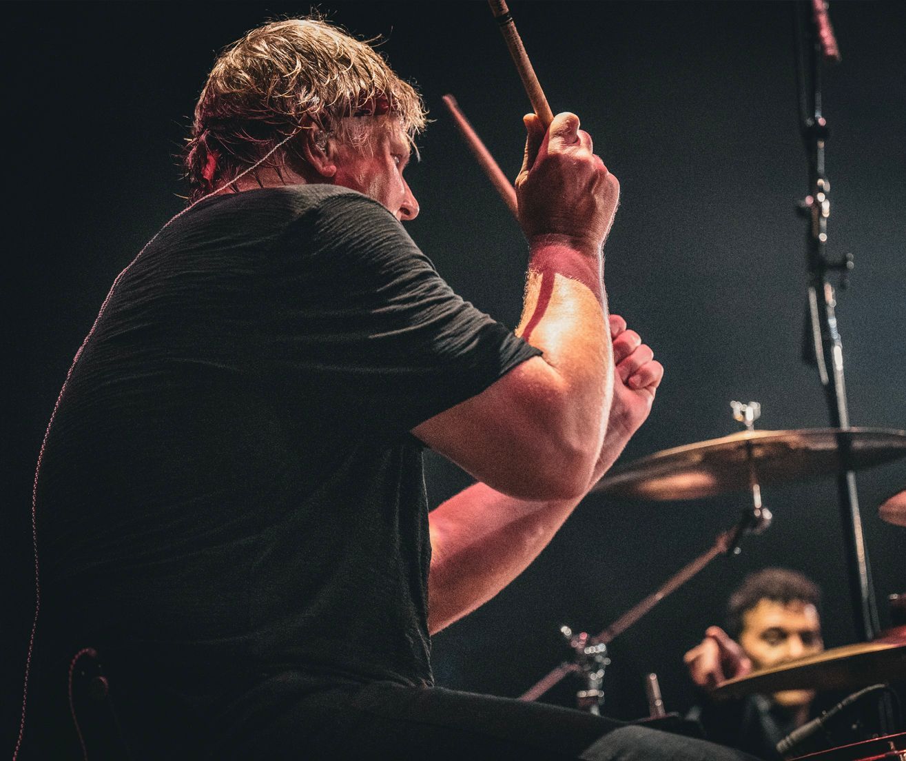 Drummer in a dark shirt playing on stage, hitting cymbals with drumsticks. Another drummer visible in background.