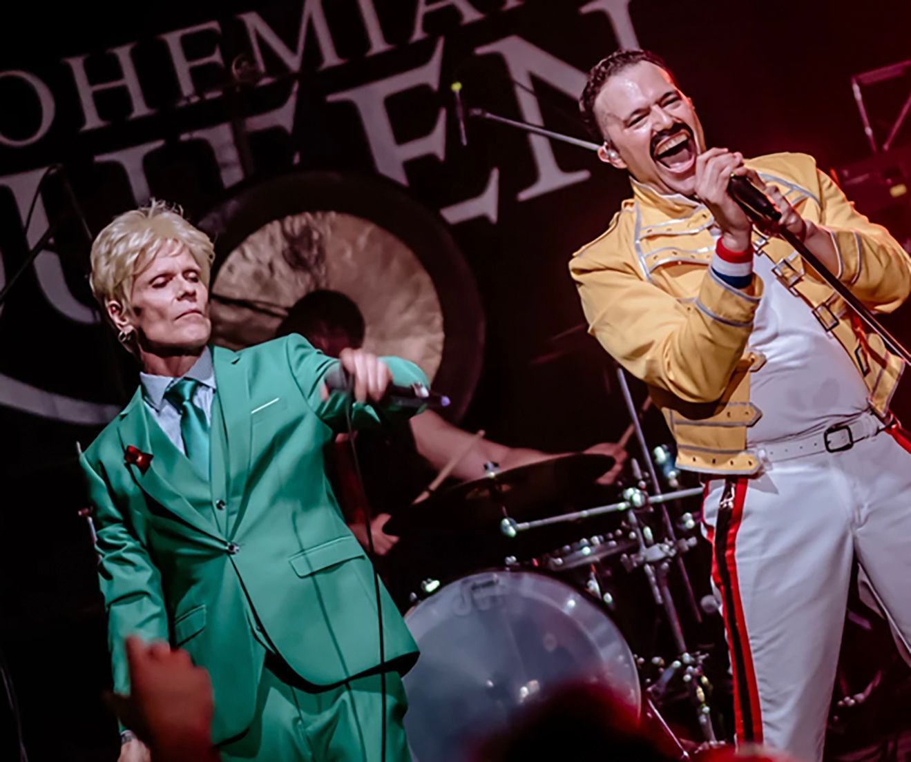 Two performers dressed as David Bowie and Freddie Mercury on stage. Bowie is in a green suit. Mercury is in a yellow jacket.