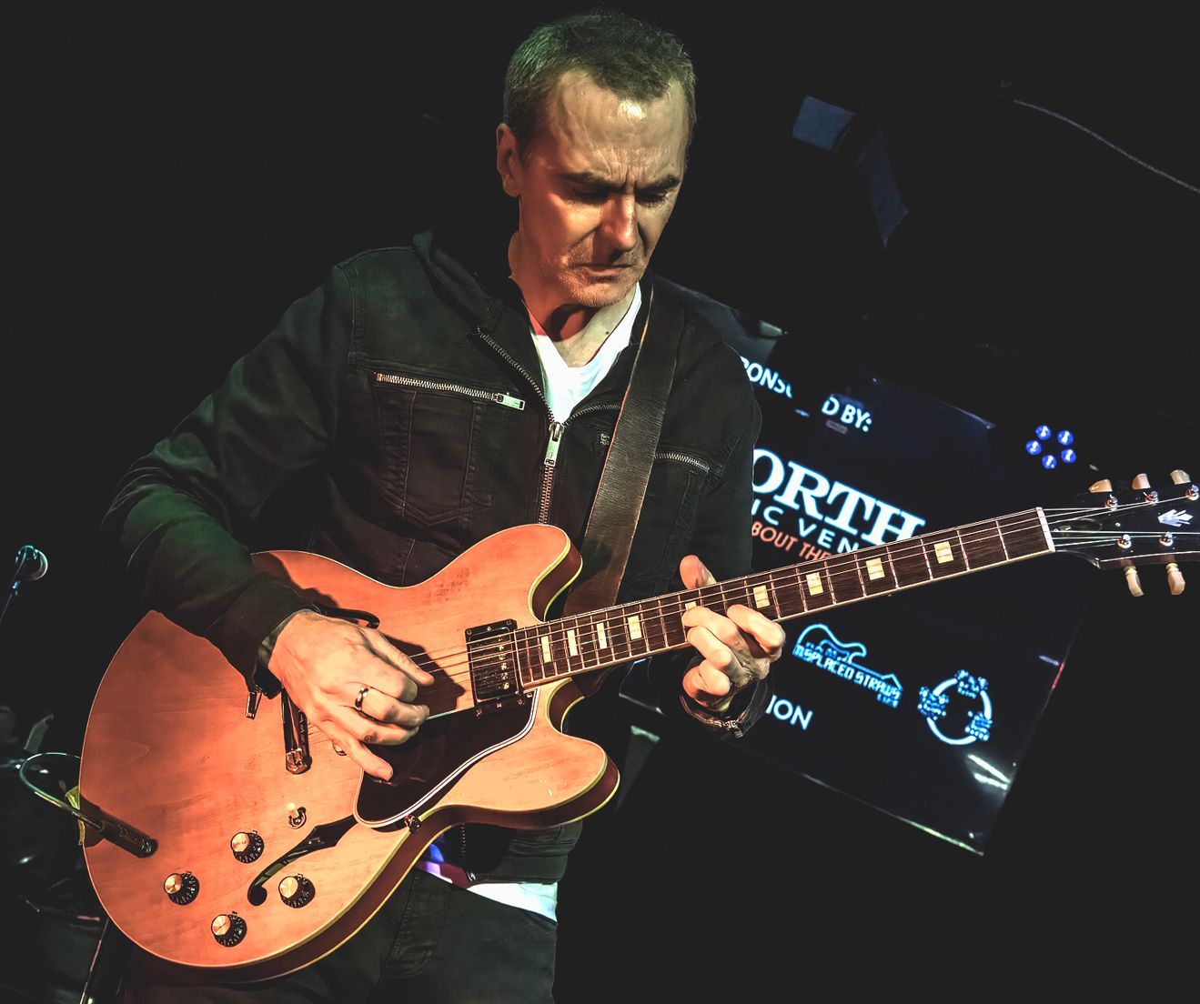Man playing a hollow body electric guitar on a stage with a dark background.