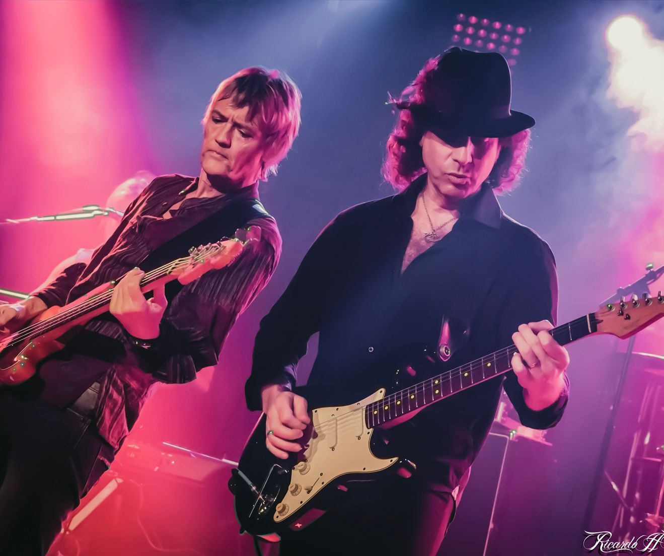 Two men are playing guitars on a stage in a dark room.