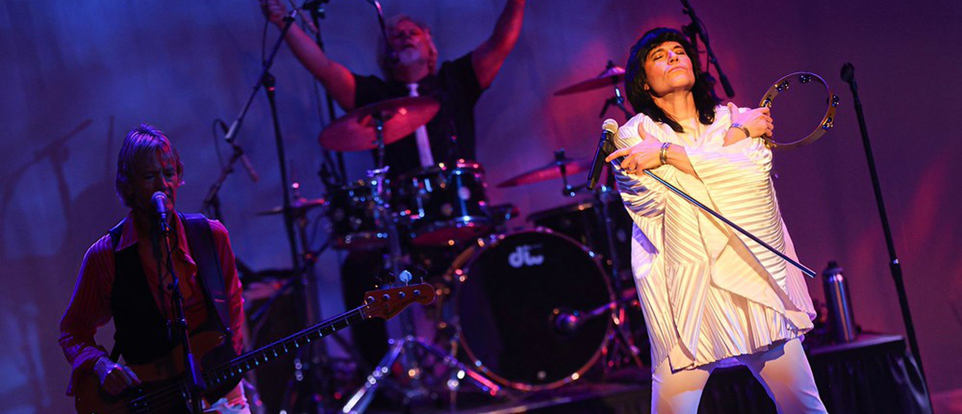 Band performing on stage: singer with tambourine, bassist, drummer with arms raised.