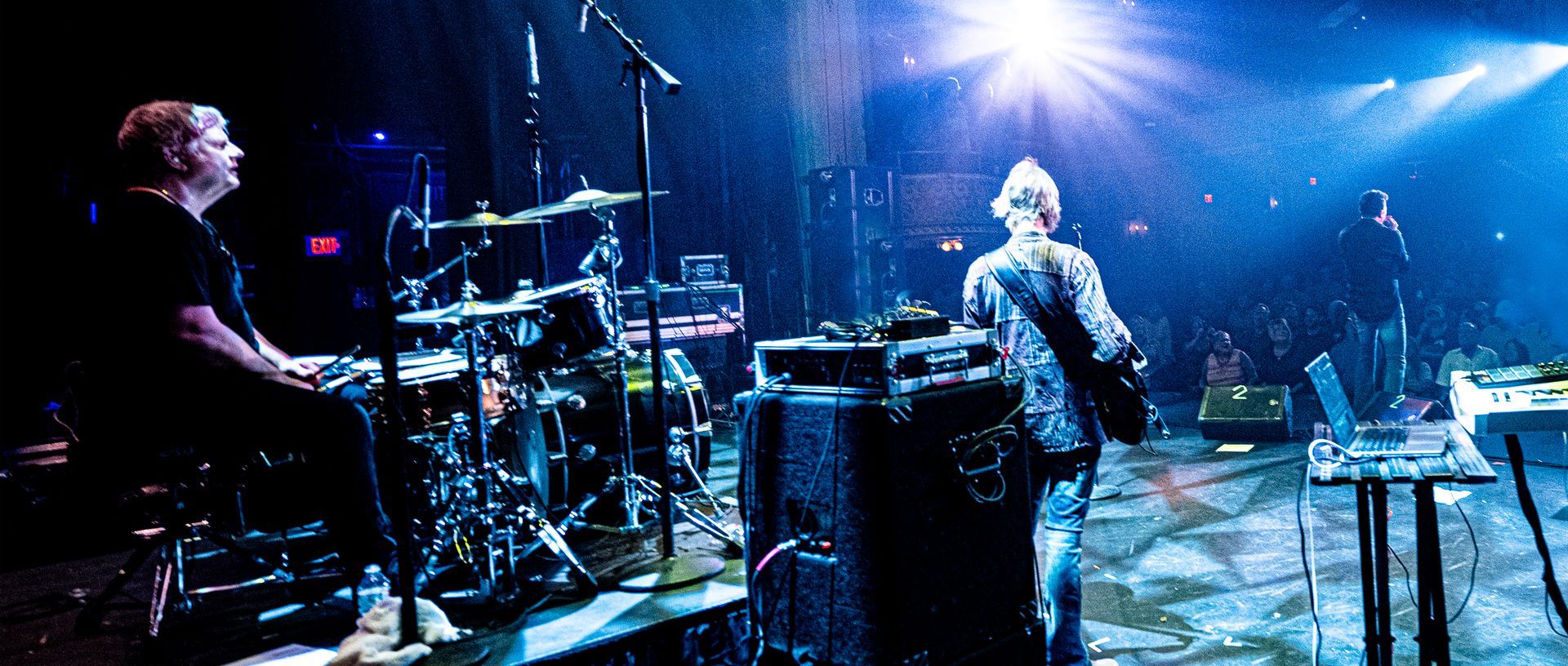 Band performing on stage, drummer on the left, other musicians facing crowd, blue stage lighting.