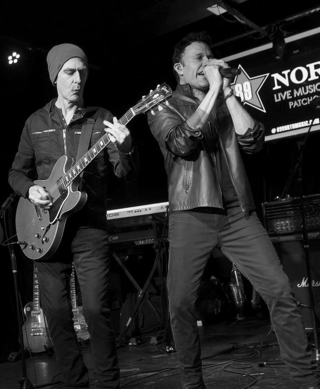 Two men on stage, one singing into a microphone, the other playing guitar. They perform at a live music venue.