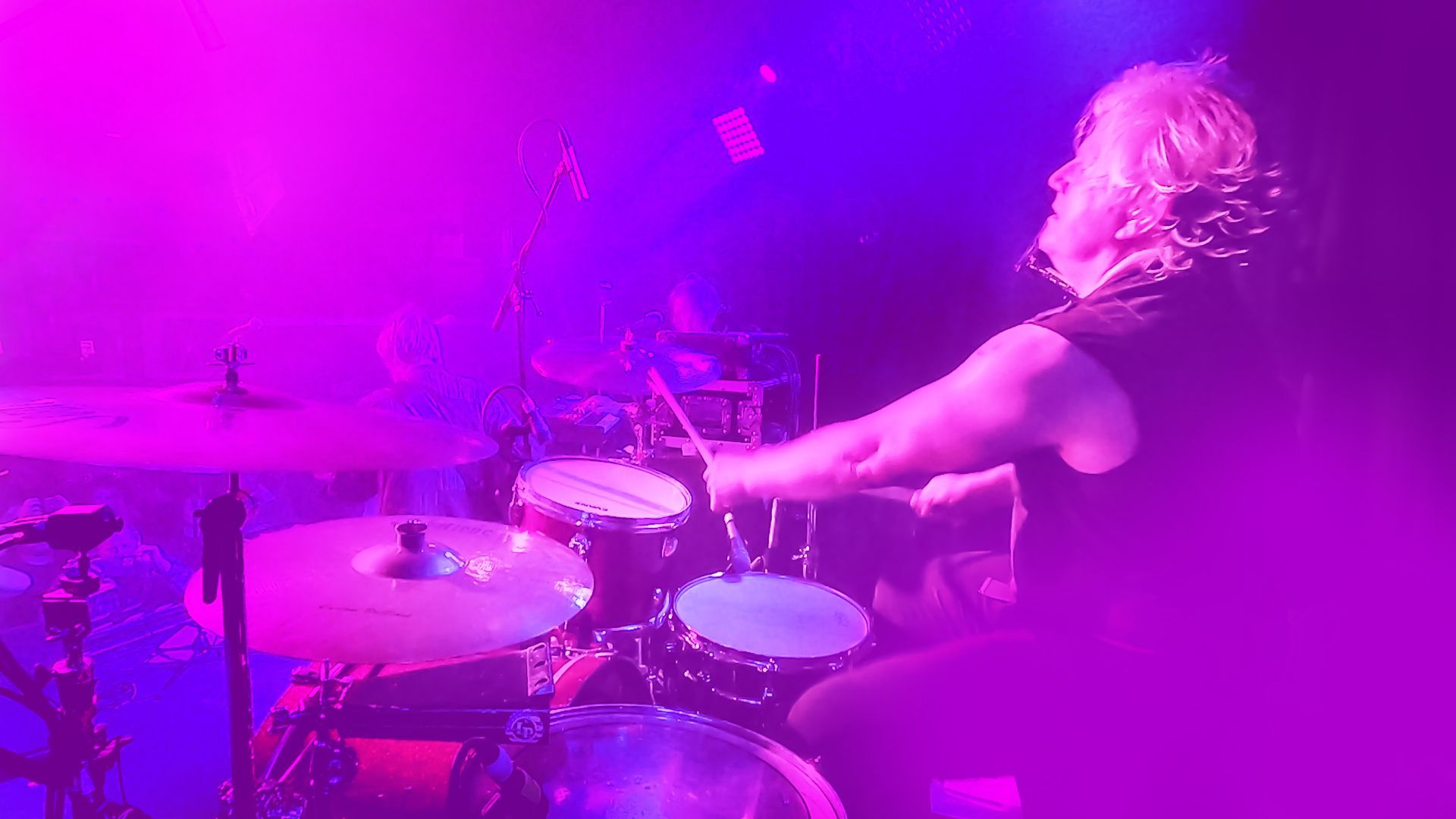 A woman is playing drums on a stage in front of a purple light.