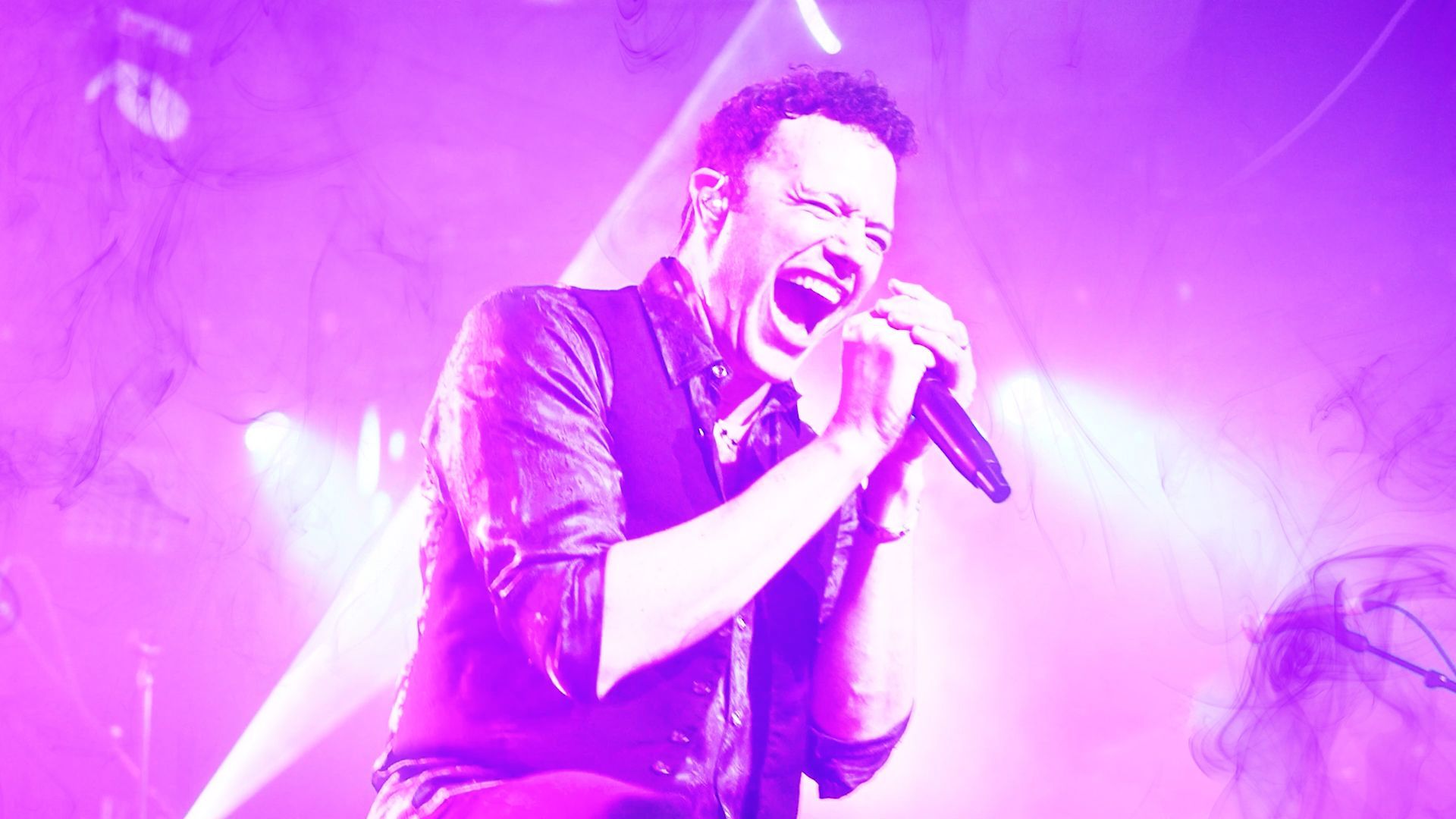 A man is singing into a microphone on a stage in front of a purple background.
