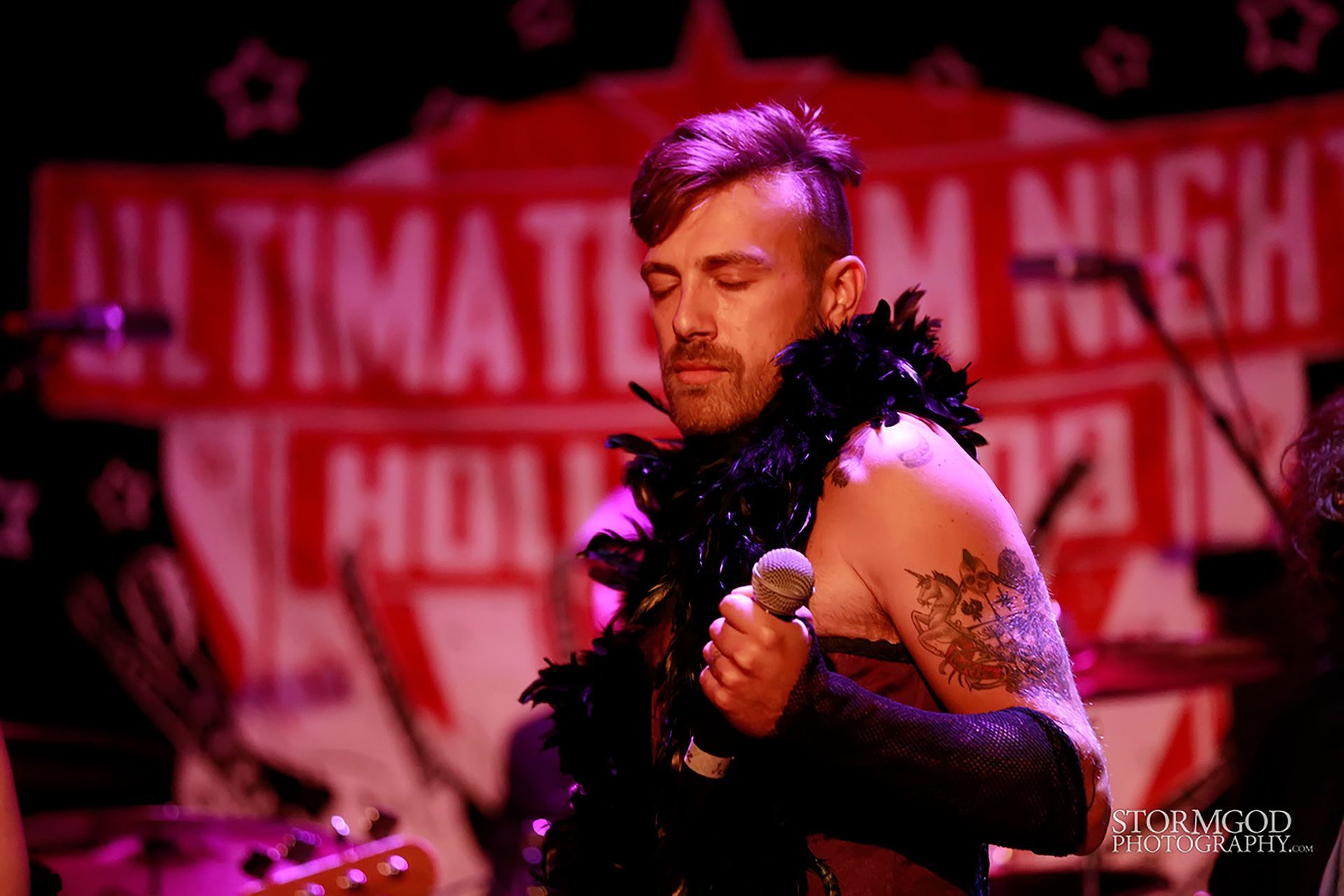 A man in a feather boa is singing into a microphone on a stage.
