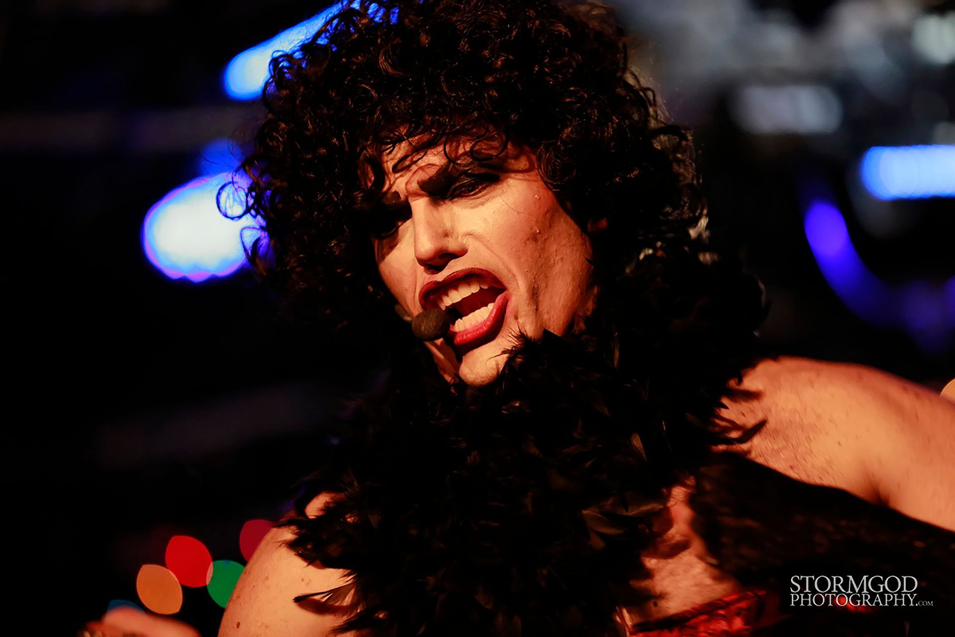 A woman with curly hair and feathers on her face is singing into a microphone.