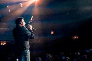 Man on stage with a microphone, lit by spotlights, gesturing to the audience. Dark setting.
