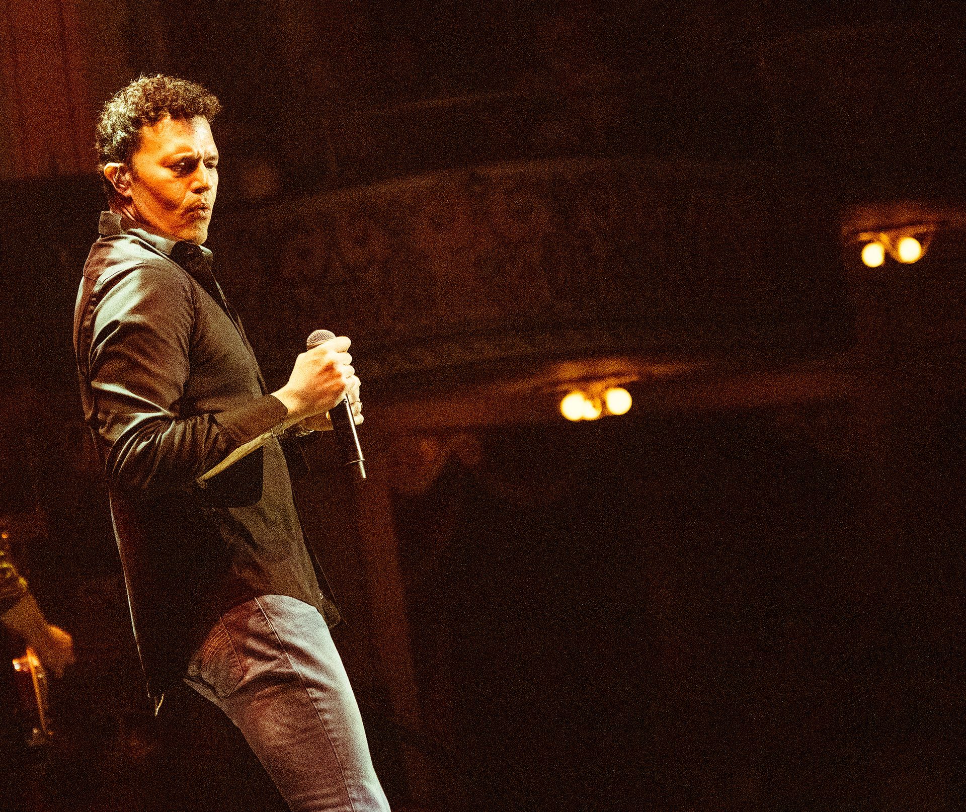 Man with microphone on stage, dressed in black shirt and jeans, singing with spotlight and two overhead lights.