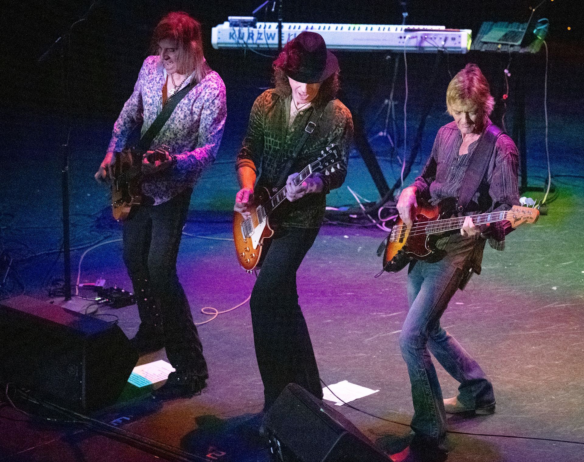 Three men are playing guitars on a stage with a keyboard in the background