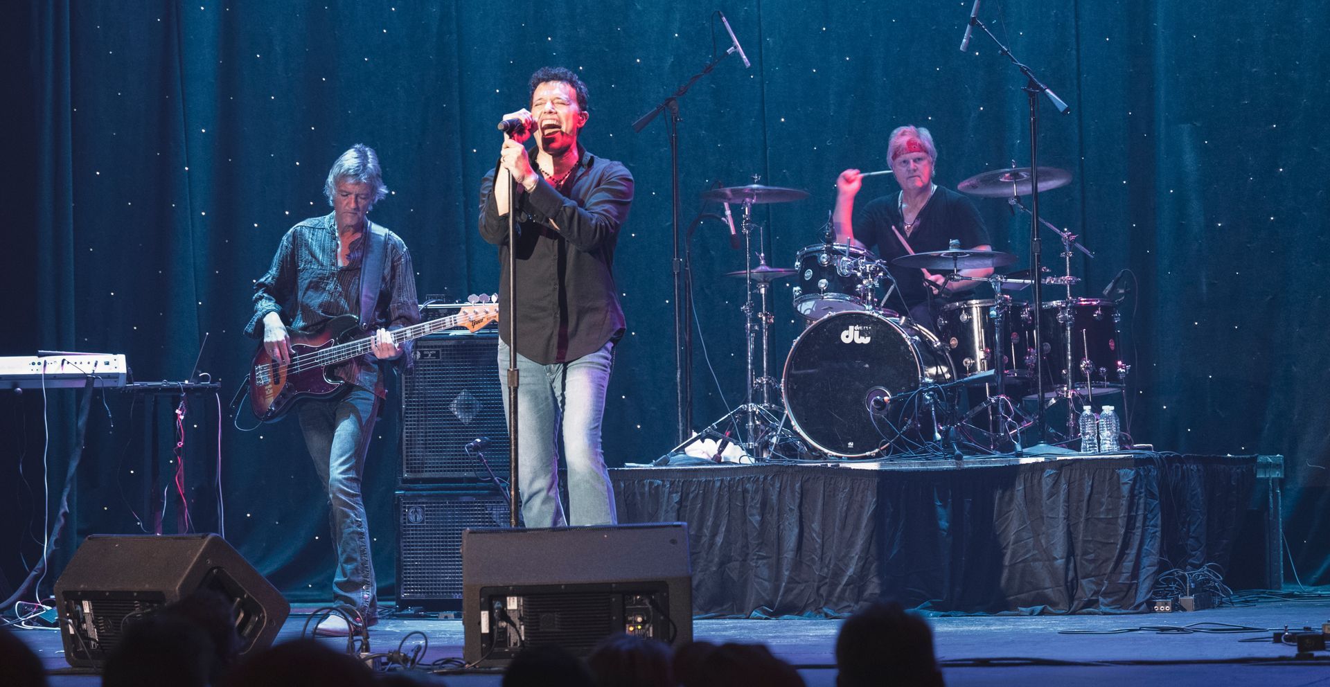 Band performing on stage: singer at center, guitarist left, drummer right, blue stage backdrop.