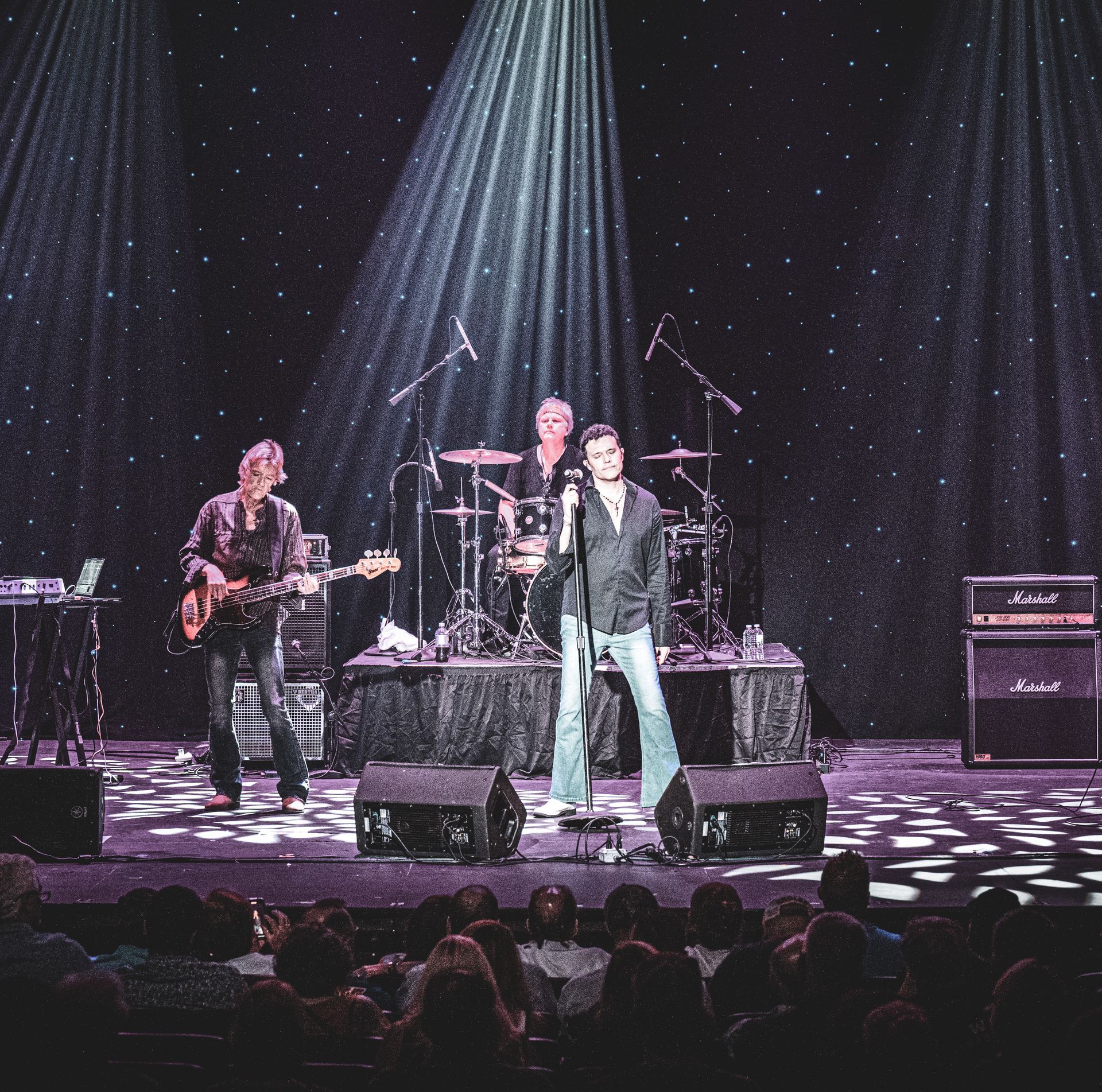 Band performing on stage, lit by spotlights. A singer with a mic, guitarist, and drummer are visible. Audience in front.