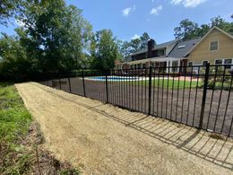 Black fence surrounds a pool and patio, gravel path in foreground, trees and house in background.