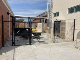 Black chain-link fence encloses a construction area near a brick building with a metal ladder.