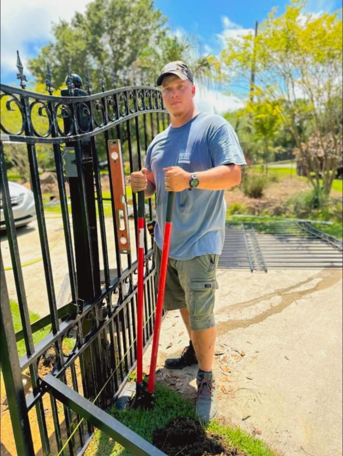Man in blue shirt and cap holding tools near a black metal gate, outdoors.