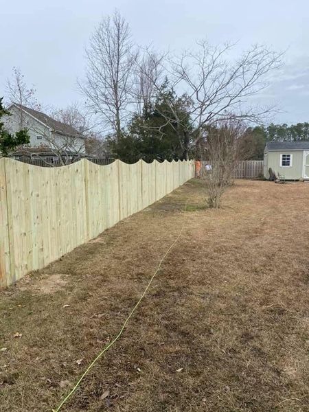 Wooden scalloped fence in a backyard with brown grass, under a cloudy sky.