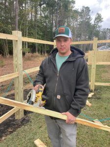 Man in gray hoodie and hat, holding a circular saw and wood, building a wooden fence outdoors.