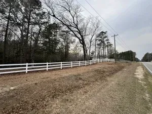 White fence lines a roadside ditch next to a wooded area and highway under a cloudy sky.