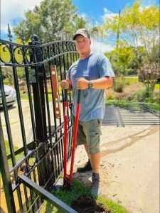 Man in cap and work clothes installing a black metal gate with tools outdoors on a sunny day.