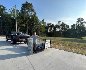 Man stands beside a truck and rolled chain-link fence on a concrete pad, with trees and open land in the background.