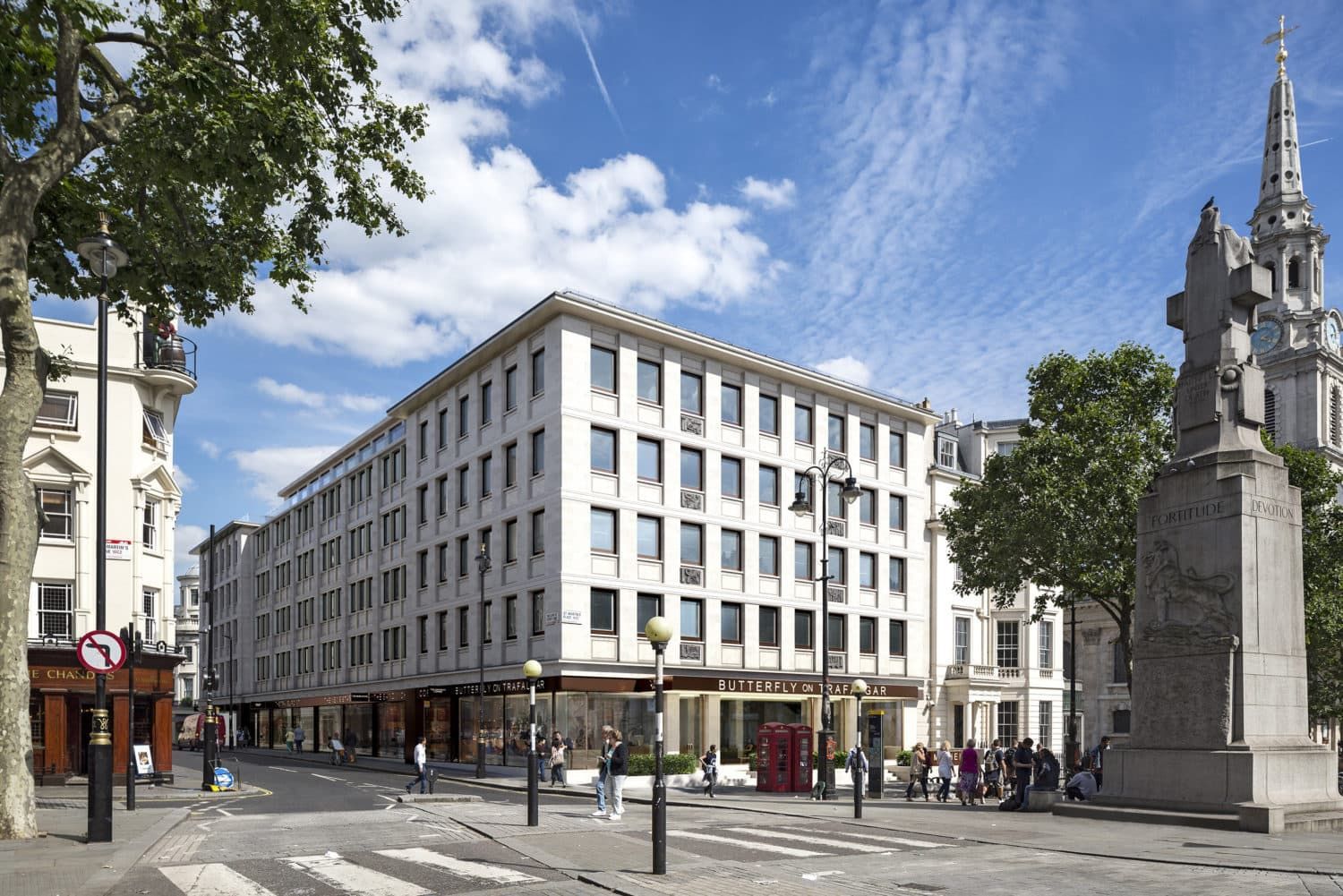 A white building with many windows, people walking on a city street, and a monument.