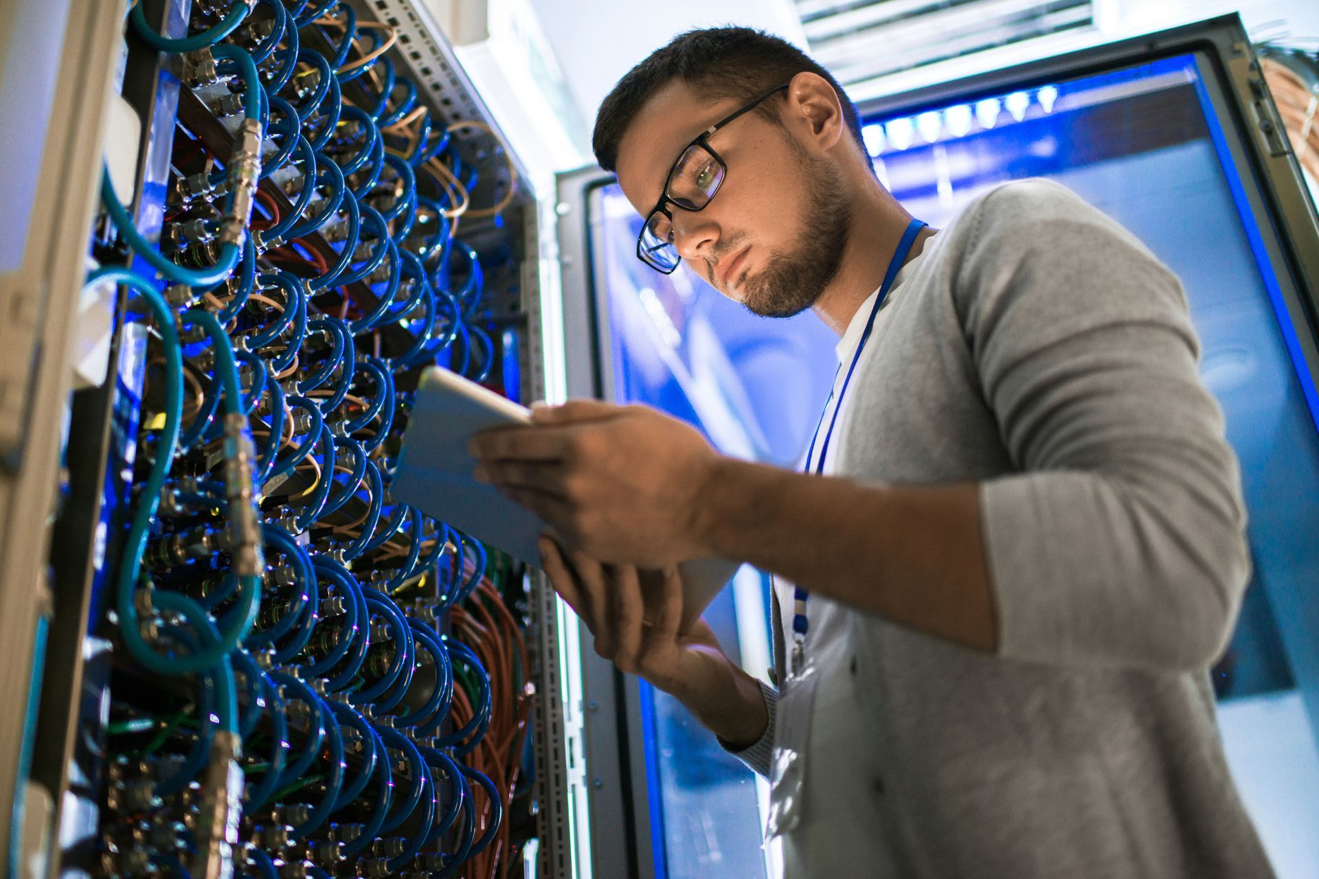Technician examining server rack with tablet; blue cables, wearing glasses in data center.