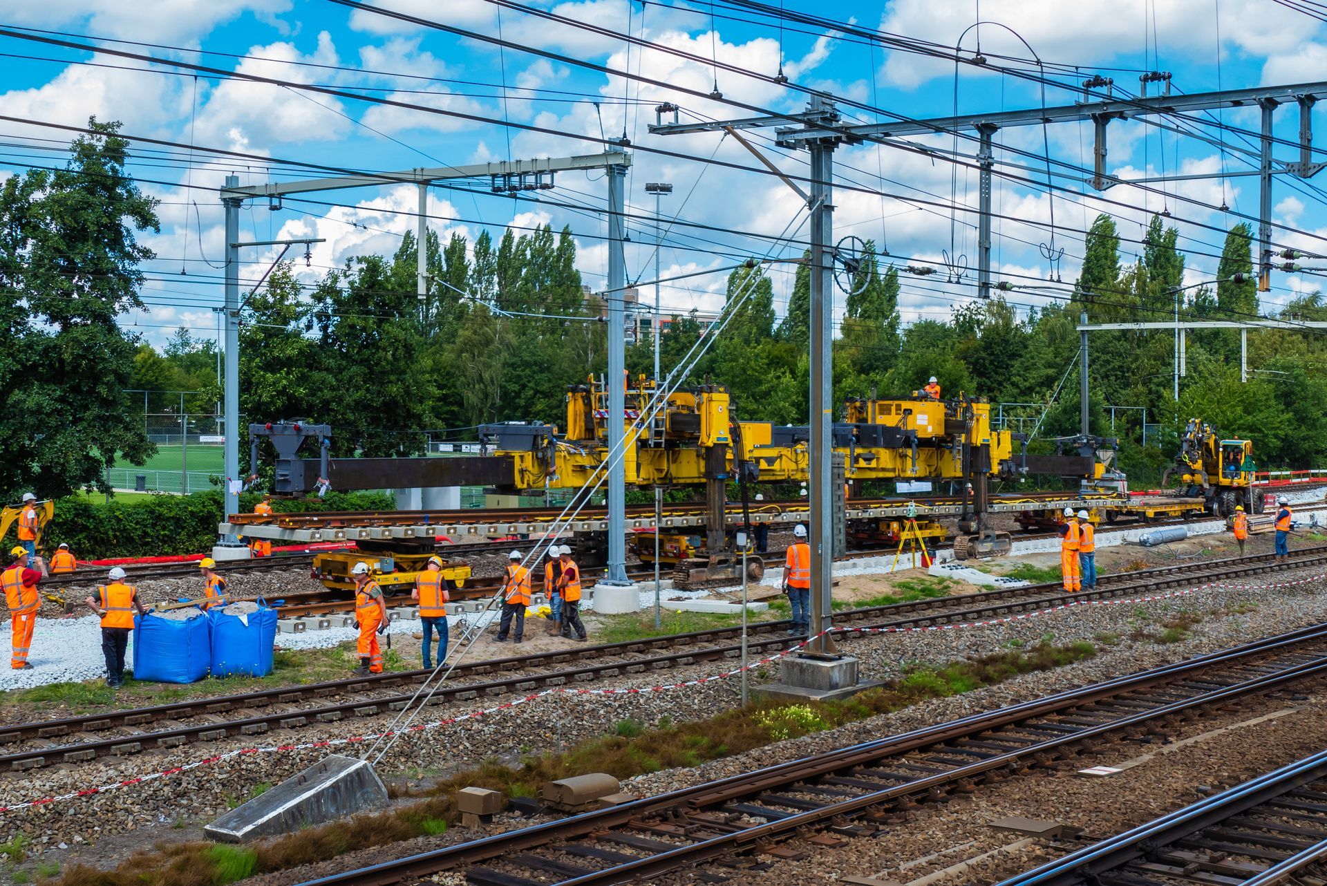 Railway track construction site with workers in orange vests, yellow machinery, overhead power lines.