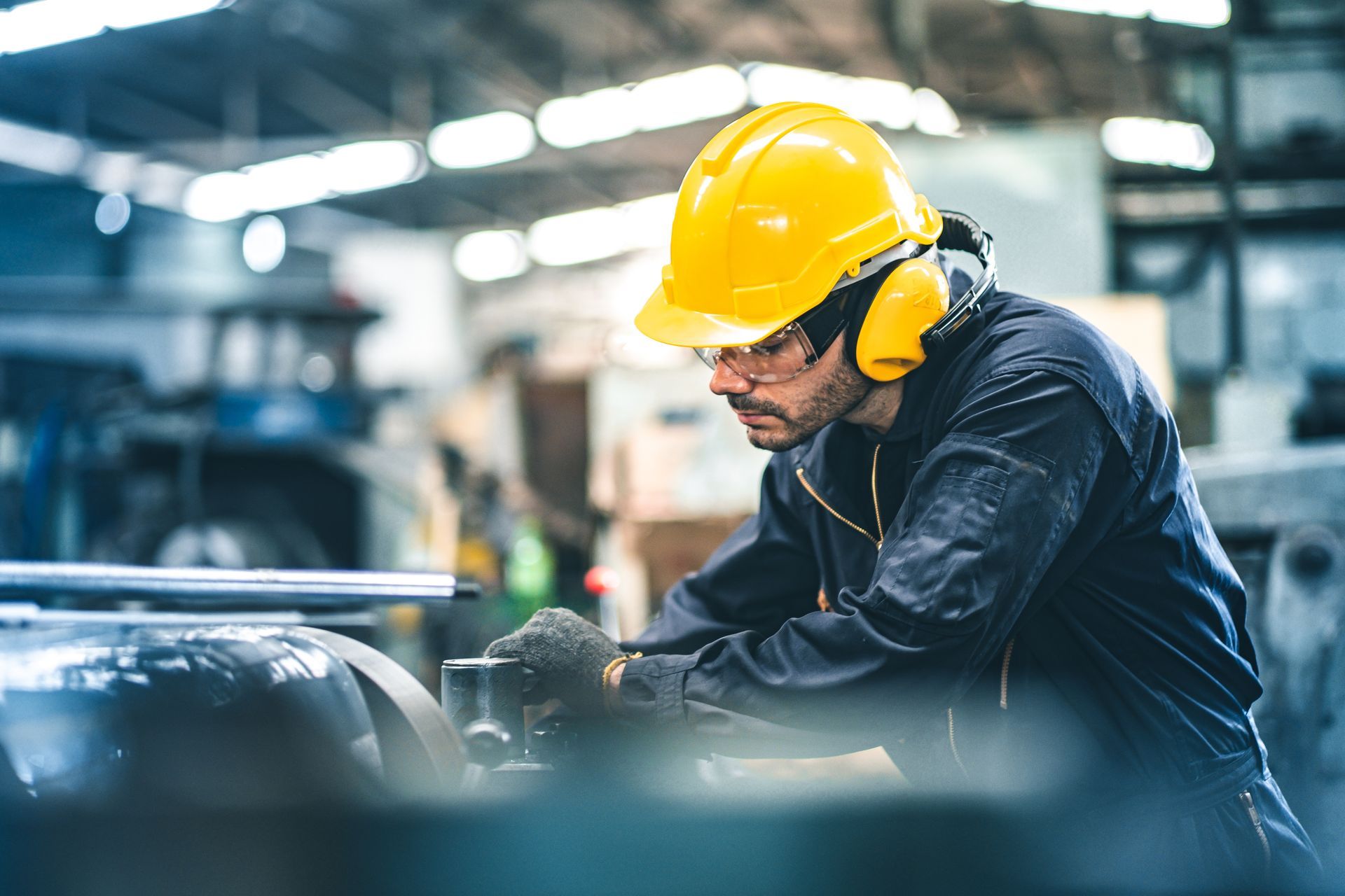 Worker in yellow hard hat and hearing protection operates machinery in a factory.