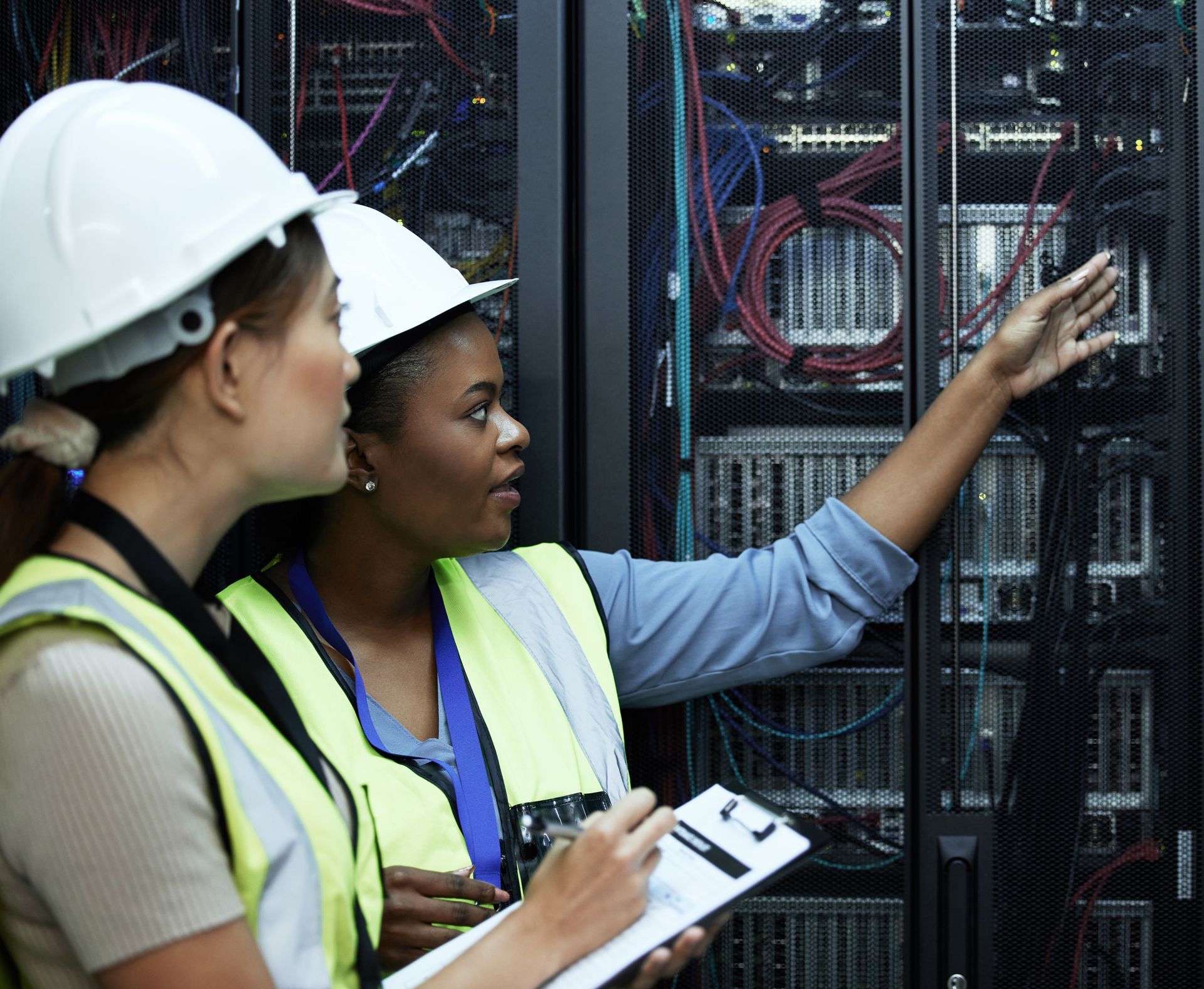 Two people in hard hats and vests examining a server rack. One points, the other holds a clipboard.