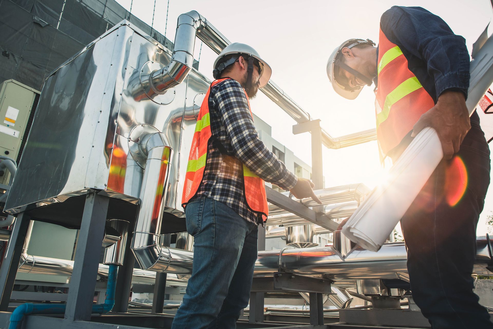 Two workers in safety vests and hard hats inspecting machinery outdoors, sunny day.