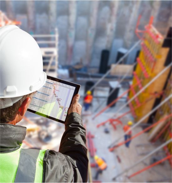 Construction worker in a white hard hat and safety vest, looking at a tablet at a construction site.