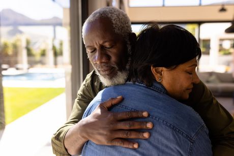 Two people embrace indoors near a sunlit window, one wearing a blue jacket.
